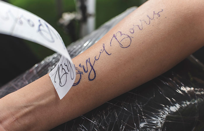 A tattoo professional removes a carbon sheet revealing the design outline of a calligraphic tattoo on a client's forearm